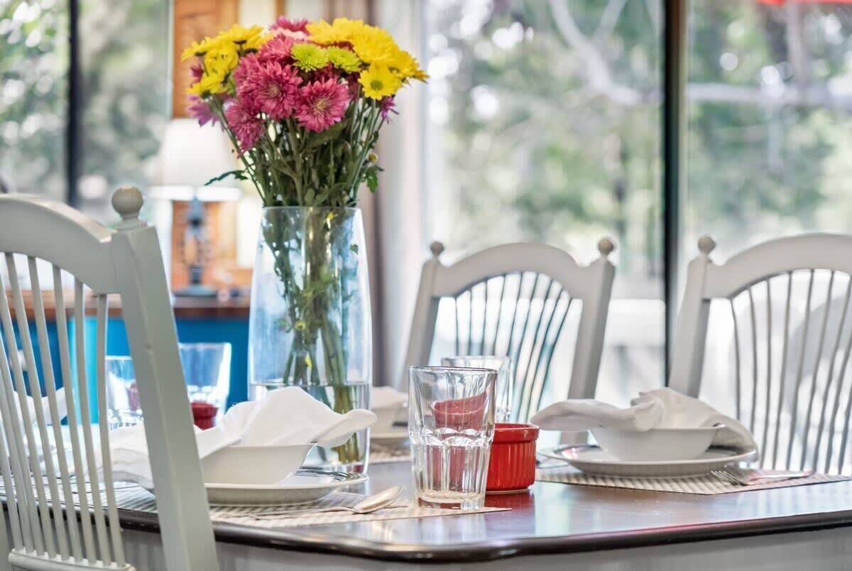 Una mesa de comedor con sillas blancas, un jarrón de flores y un vaso de agua.
