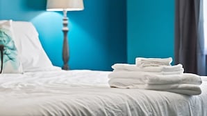 A neatly made bed with white linens, a stack of folded white towels, and a lamp on a bedside table in a room with teal walls. - Eastview Drive Jolly Green Giant (Madison)