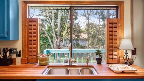 A kitchen with a wooden countertop, a double sink, and a view of a backyard with trees and a deck. - Eastview Drive Jolly Green Giant (Madison)