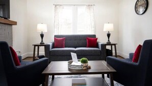 A living room with a grey sofa, red cushions, two side tables with lamps, a coffee table with a laptop and a small plant, and a clock on the wall. - 28th Street Historic at Norwood (Birmingham)