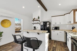 A modern kitchen with white cabinetry, a central island with a marble countertop, and a built-in oven. There are two black bar stools and a potted plant. - Byrd 4 Tiny Stay Getaway (Owens Cross Roads)