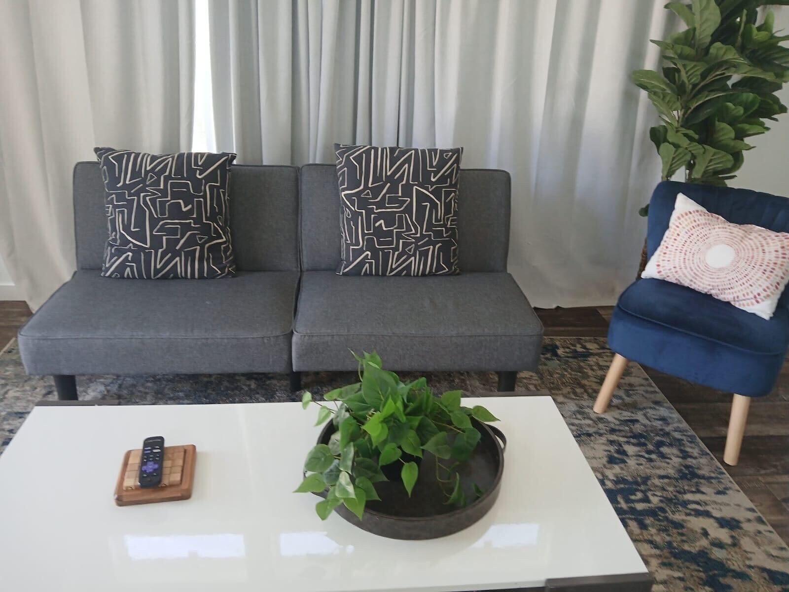 Un salon moderne avec un canapé gris, deux coussins à motifs, un fauteuil bleu, une table basse blanche avec une télécommande et une plante en pot.