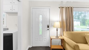 Une pièce avec une porte blanche, une petite table avec une lampe, un canapé beige et une vue sur la verdure à l’extérieur.