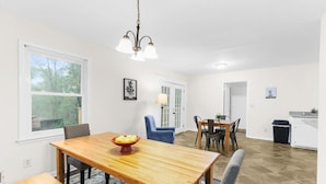 Un coin repas avec une table en bois, des chaises et un lustre. Il y a une fenêtre donnant sur des arbres et une porte menant à une autre pièce.
