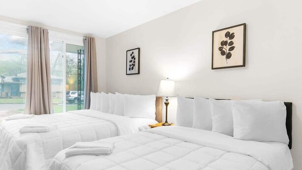 Une chambre d’hôtel avec deux lits, une grande fenêtre avec des rideaux, une table de chevet avec une lampe et des œuvres d’art encadrées accrochées aux murs.