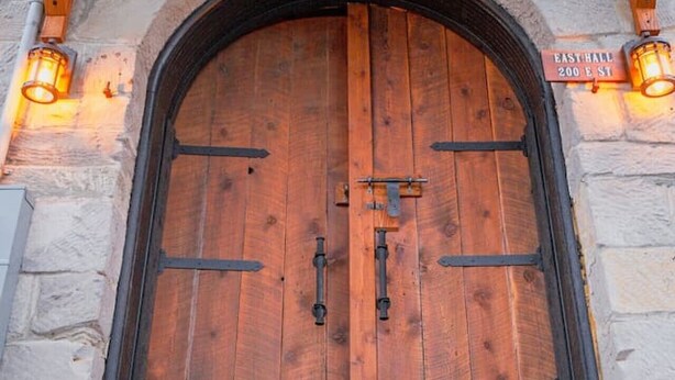 Une porte en bois en forme d’arc, dotée d’un loquet métallique, flanquée de murs en pierre et de poutres en bois, et située sous un porche abrité.