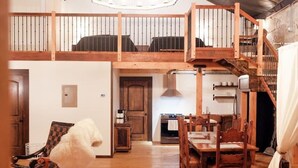 A spacious loft-style room with a high ceiling, wooden beams, a dining area with a table and chairs, a kitchen area, and a staircase leading to an upper level. - The Scandinavian West Hall—Near Anthony Lakes! (North Powder)