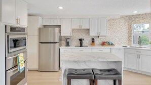 A modern kitchen with white cabinets, a stainless steel refrigerator, a marble countertop, and a window with a view of greenery. - Coastal Oasis ~  Beaches, Parks, Shopping & Entertainment, Pet Friendly (Venice)