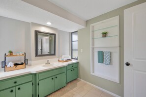 A modern bathroom with a large mirror, green cabinets, a white sink, and a glass shelf with towels.