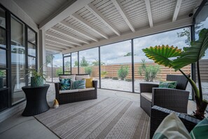 Een overdekt terras met rieten meubels, een grote potplant en uitzicht op de achtertuin.