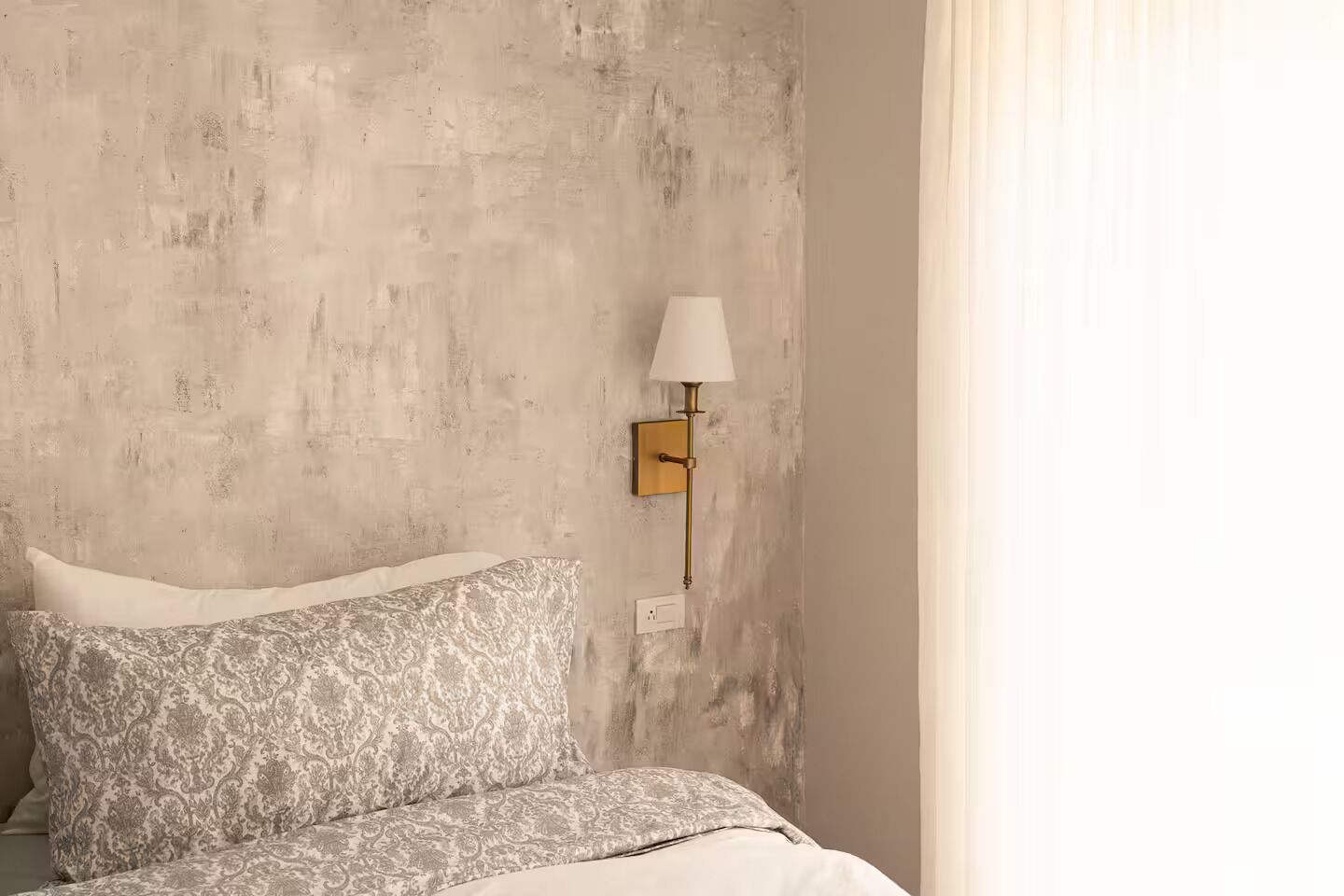 A bedroom with a bed, patterned pillows, a textured wall, and a wall-mounted lamp.