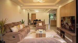 Ein modernes Wohnzimmer mit einem braunen Sofa, einem Couchtisch und einem Fernseher auf einem Holzboden. Im Hintergrund befindet sich ein Essbereich mit Tisch und Stühlen.