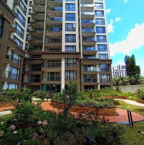 Um edifício residencial com vários andares e varandas, cercado por jardins ajardinados e um caminho pavimentado.