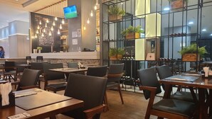 Ein Restaurantinneres mit Holztischen und -stühlen, einer Bar mit Fernseher und einem großen Fenster mit Pflanzen.