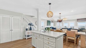 A spacious kitchen with a central island, white cabinetry, and a dining area with wooden chairs and a table. - Just A Touch by Brunswick Vacation Rentals: 6BR Oceanfront Retreat, Sleeps 16 (Ocean Isle Beach)