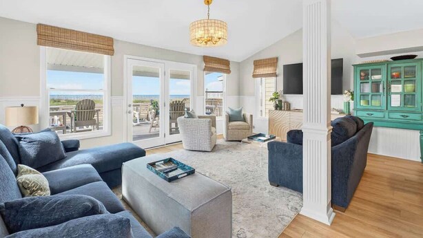 A spacious living room with a large window, a chandelier, and a teal cabinet. - Just A Touch by Brunswick Vacation Rentals: 6BR Oceanfront Retreat, Sleeps 16 (Ocean Isle Beach)