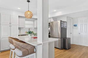 Cozinha moderna com armários brancos, geladeira de aço inoxidável, uma pequena ilha com banquetas e luminária pendente.