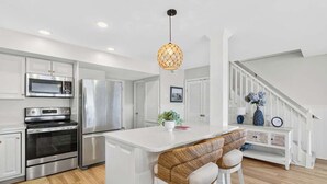 A kitchen with stainless steel appliances, a white island with bar stools, and a staircase with white railings. - Just A Touch by Brunswick Vacation Rentals: 6BR Oceanfront Retreat, Sleeps 16 (Ocean Isle Beach)