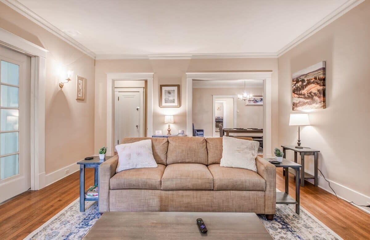 Una sala de estar con un sofá color beige, una mesa de centro y una alfombra. Hay dos mesitas de noche con lámparas, un cuadro enmarcado en la pared y una puerta que da a otra habitación.