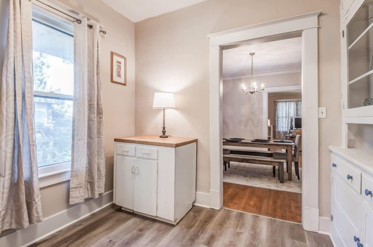 Una habitación con piso de madera, un armario blanco con cajones, una mesa con sillas y una araña.
