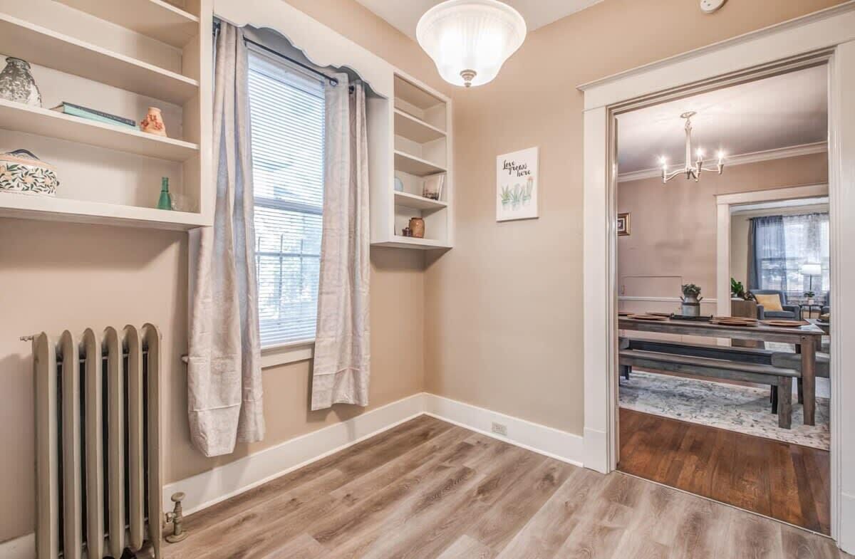 A room with a wooden floor, a radiator, a window with curtains, a shelf with decorative items, and a door leading to another room.