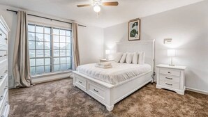 Un dormitorio con una cama grande, un ventilador de techo, una ventana con cortinas, una mesita de noche con una lámpara y un aparador.