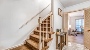Una escalera con peldaños alfombrados y balaustres blancos, que conduce a una habitación con un escritorio y una silla, una ventana y una puerta con paneles de celosía.