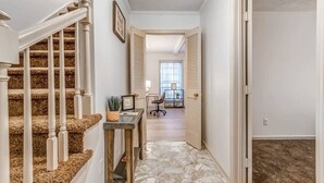 Un pasillo con escaleras cubiertas de alfombra, una pequeña mesa con una planta, y vista a una habitación con ventana y sillón.