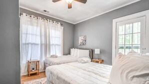 Ein Schlafzimmer mit zwei Betten, einem Deckenventilator, einem Fenster mit Vorhängen, einem Nachttisch mit Lampe und einem Teppich auf dem Boden.
