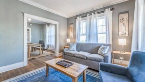 Ein Wohnzimmer mit grauem Sofa, blauem Sessel und einem Holztisch. Im Hintergrund steht ein Esstisch mit Stühlen.
