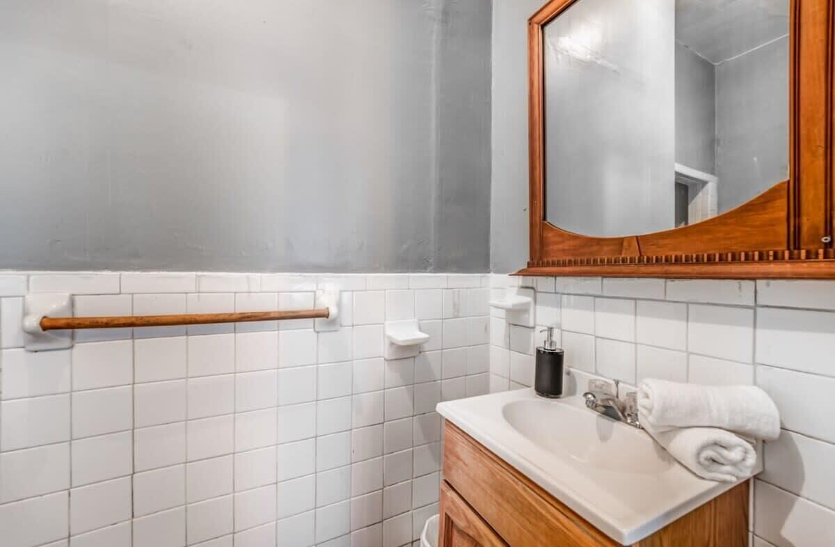 Un bagno con pareti piastrellate di bianco, un mobile lavabo in legno, uno specchio, un asciugamano e un dispenser per sapone.
