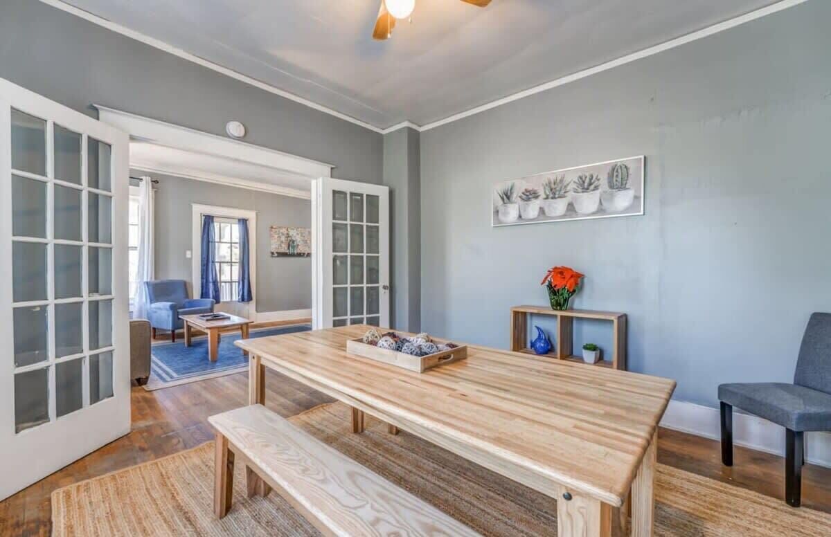 Une pièce spacieuse avec une table à manger en bois, des bancs et un tapis bleu. On y trouve une chaise et une petite table sur laquelle est posé un vase de fleurs.