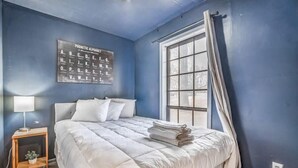 Una camera da letto con un letto, un comodino, una lampada, una finestra con tende e un poster con l'alfabeto fonetico appeso al muro.