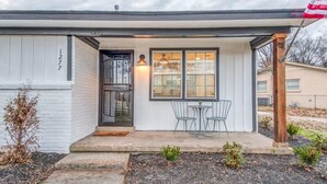 Een gelijkvloers huis met een witte gevel, nummer 1277, voorzien van een voortuin met twee stoelen en een tafel, een kleine tuin en een houten paal met een Amerikaanse vlag.
