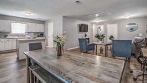 A modern kitchen with white cabinets, a dining area with a wooden table, blue chairs, and a TV mounted on the wall.