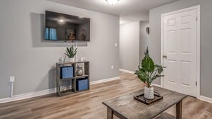 A modern living room with a flat-screen TV mounted on the wall, a wooden coffee table with a plant, and a door to the right.