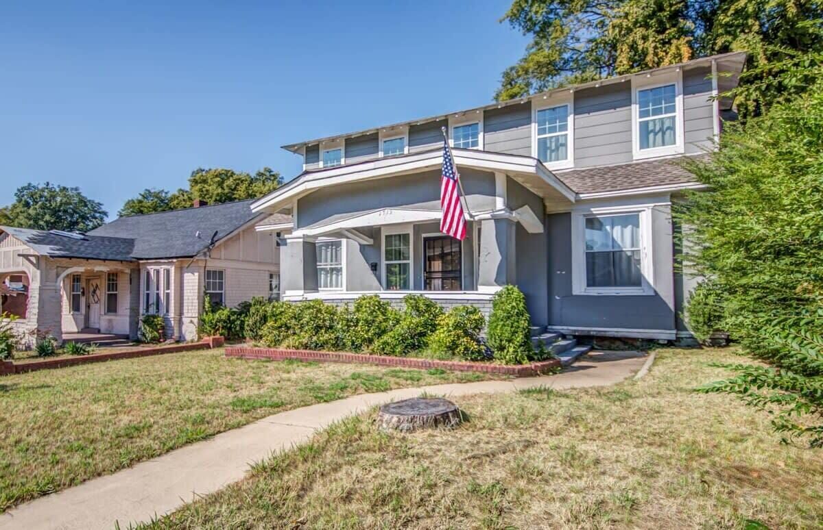 A two-story house with a front porch, a small garden, and an American flag.
