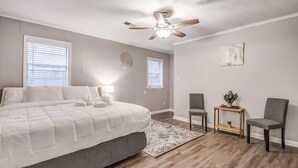 A bedroom with a bed, two chairs, a small table with a plant, a ceiling fan, and two windows with blinds. - Flamingo Hideaway (Memphis)