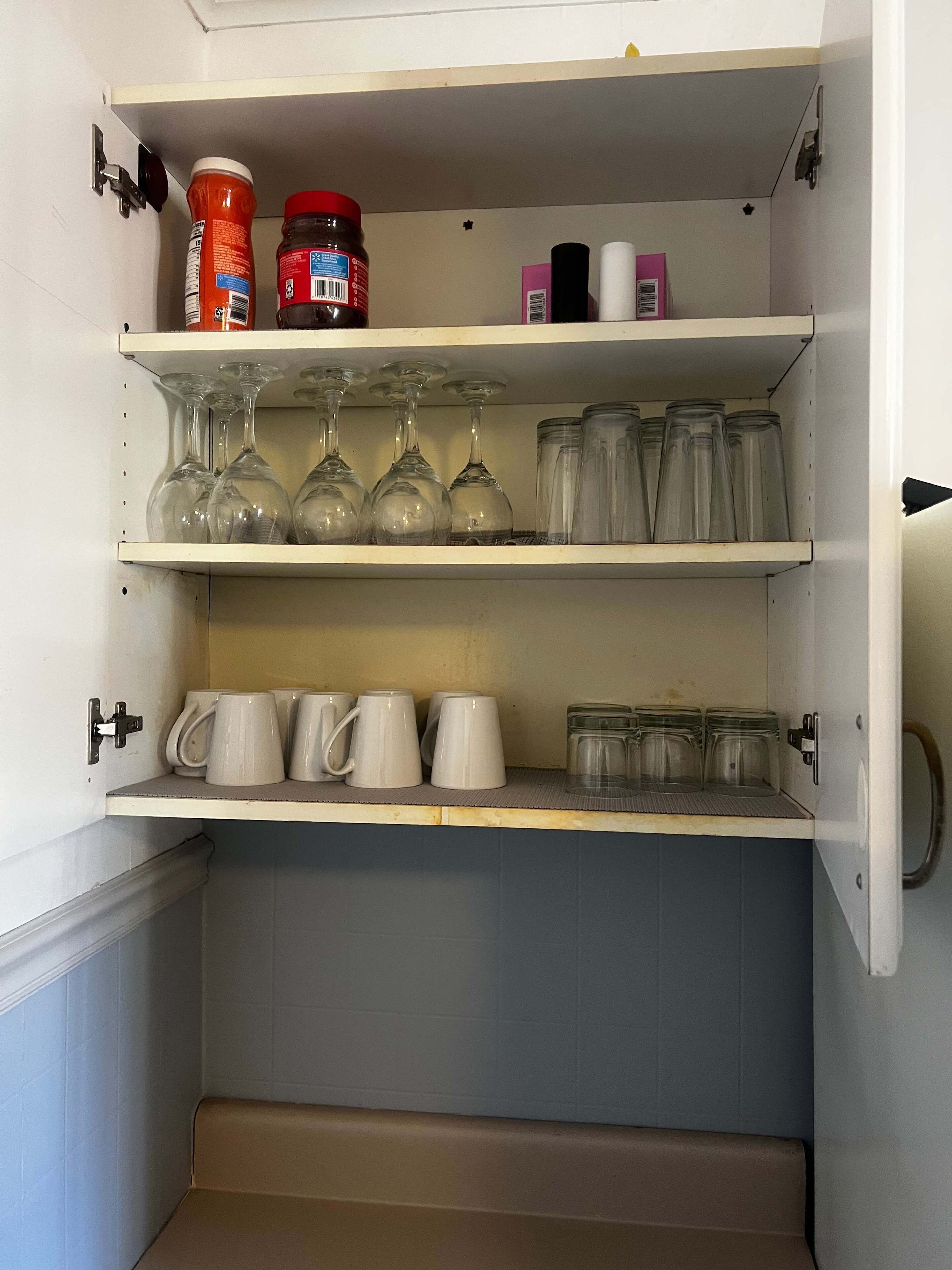 A kitchen cabinet with shelves holding wine glasses, mugs, and jars.