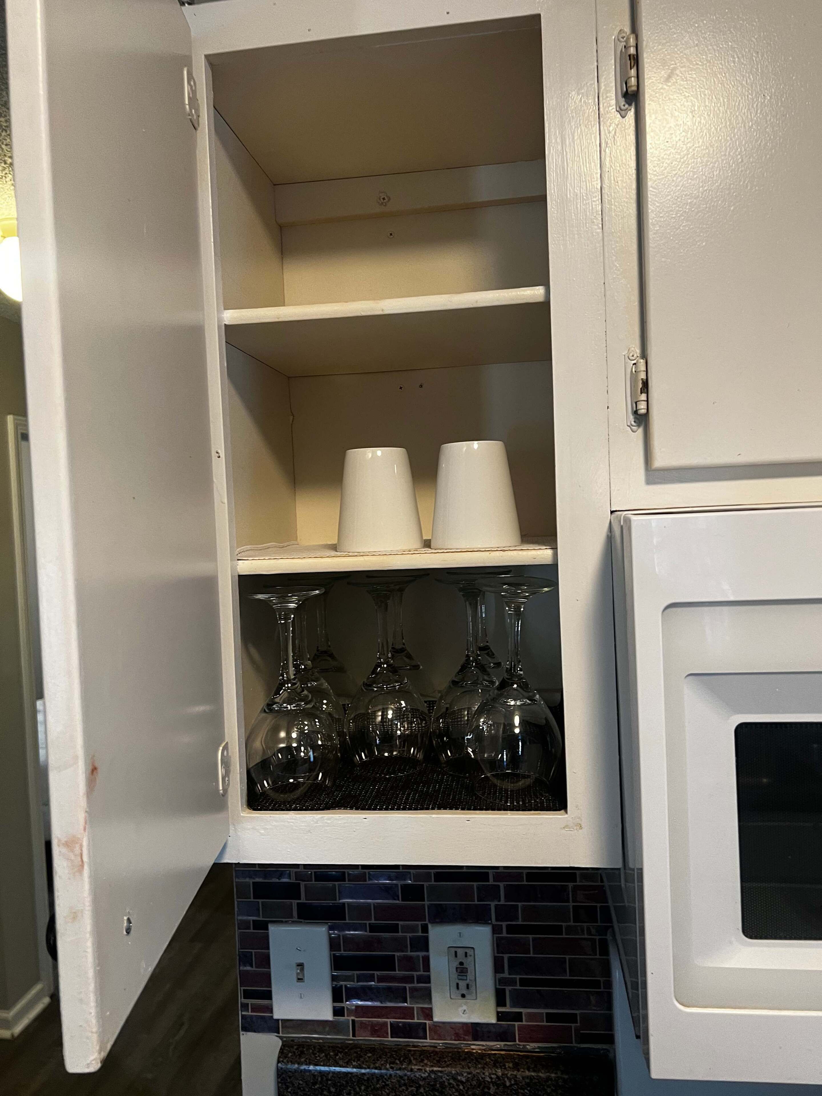 A kitchen cabinet with two shelves, one holding white cups and the other holding wine glasses.