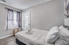 A bedroom with a bed, a window with sheer curtains, a door, and a small table with a lamp.