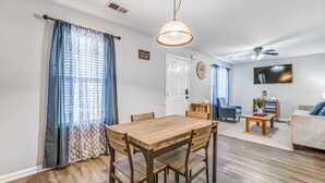 A living room with a wooden dining table, blue curtains, a ceiling fan, and a TV mounted on the wall. - Cedar Ave Escape (Memphis)