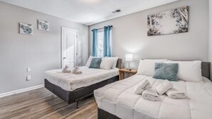 A bedroom with two beds, a window with curtains, a wall-mounted artwork, and a bedside table with a lamp. - Cedar Ave Escape (Memphis)