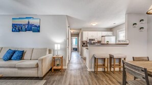 A modern living room with a sofa, a coffee table, and a dining area with a wooden table and chairs. There is a kitchen visible in the background with white cabinets and a sign that reads "Our Kitchen". - Cedar Ave Escape (Memphis)