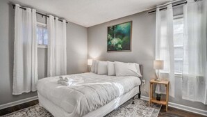 A bedroom with a large bed, white curtains, a painting on the wall, and a small bedside table with a lamp. - Yale Gather Hideaway (Memphis)