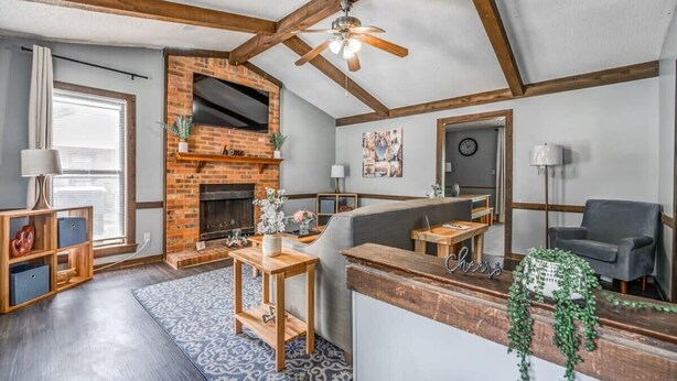 Een gezellige woonkamer met zichtbare houten balken, een open haard van baksteen, een plafondventilator en een moderne zithoek.