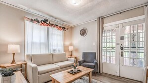 Ein Wohnzimmer mit Sofa, Sesseln, Couchtisch und Lampen. Eine Tür mit Glasfeldern und eine Uhr an der Wand sind ebenfalls zu sehen.