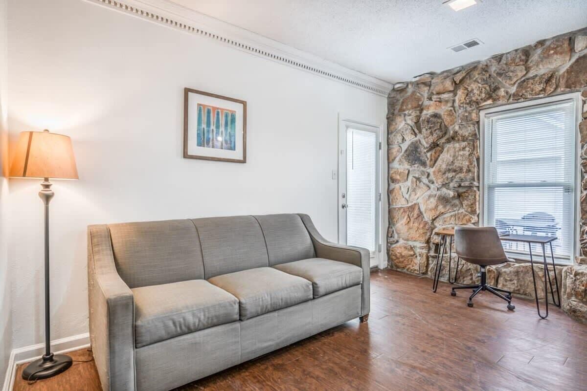 Un salon avec un canapé gris, une lampe sur pied, un mur décoratif en pierre, un tableau encadré et une fenêtre avec des stores.