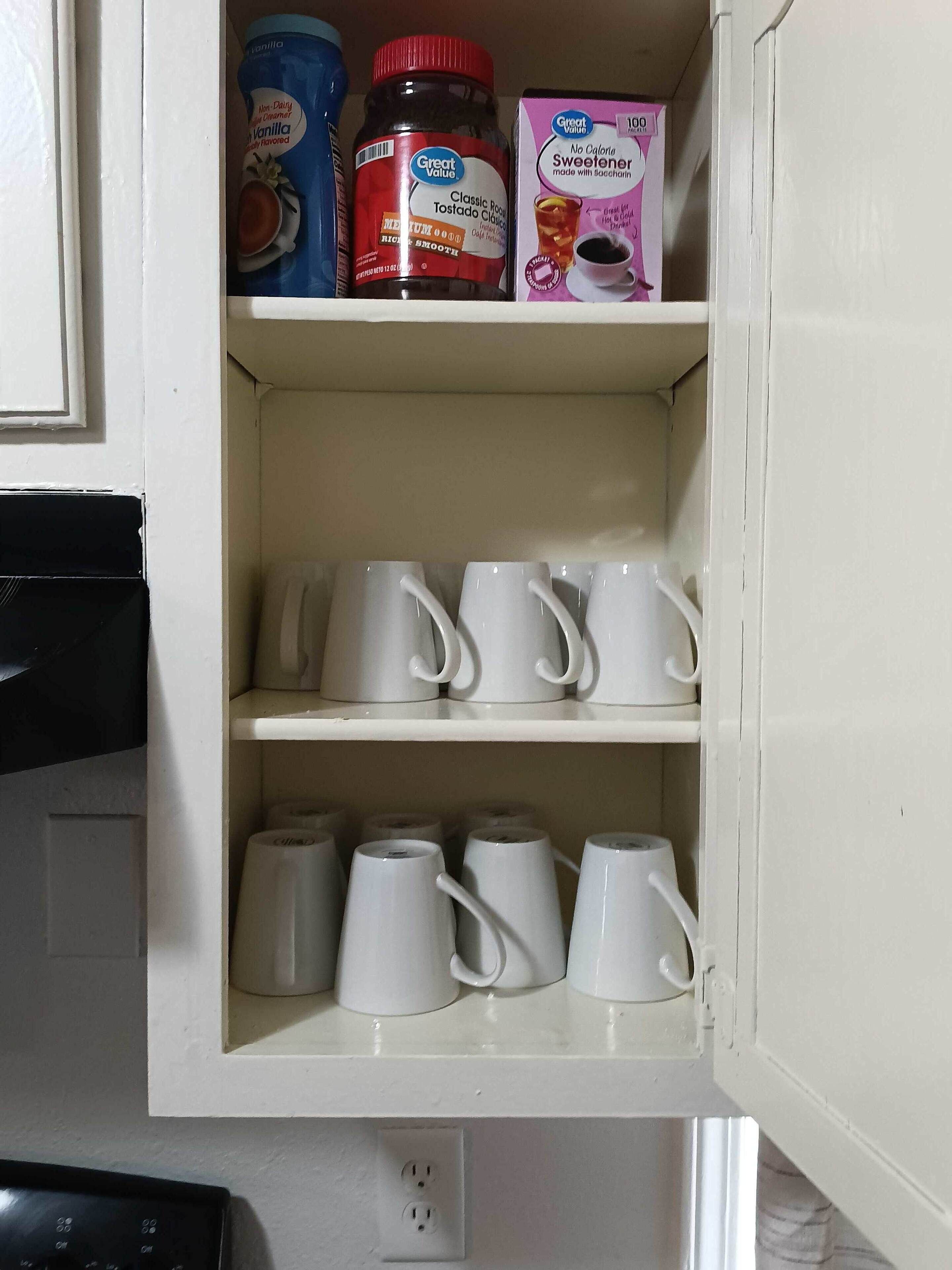 A kitchen cabinet with a white interior, containing a variety of kitchen items including a jar of vanilla-flavored creamer, a bottle of non-dairy creamer, a box of sweetener, and several white mugs and teapots.