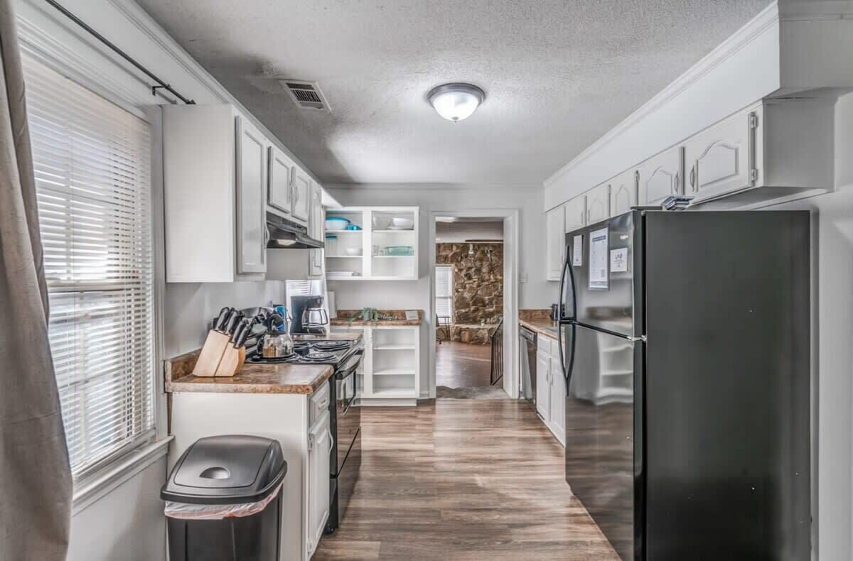 A narrow kitchen with white cabinets, a black refrigerator, and a window with blinds.